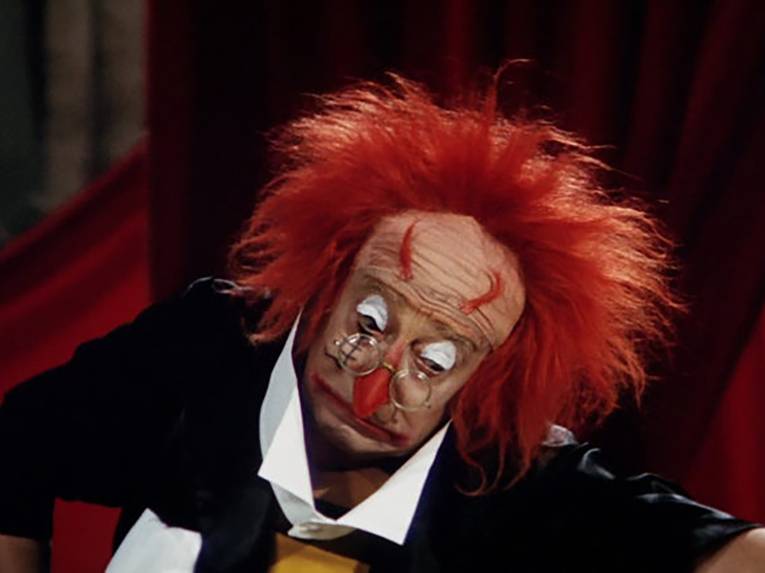 Auf dem Foto ist ein Clown  zu sehen. Er hat eine rote Strubbelmähne, die Augenlider sind weiß geschminkt und an den Augenbrauen kleben Haarbüschel, die an Teufelshörner erinnern. Auf der rot geschminkten langen Nase sitzt eine runde Brille. Der rot geschminkte Mund bildet einen graden Strich.